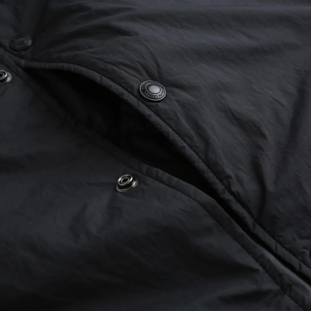 P650 Ralph Lauren pony embroidered light cotton jacket. The fabric is made of high-density nylon taffeta fabric, which combines pragmatism and classic texture. The density is about 210T-300T, the surface is smooth and slightly matte, it has excellent windproof and waterproof properties (with DWR water-repellent treatment), it has strong wrinkle resistance and is easy to care for, which is in line with the positioning of American casual "durable and practical". Classic dark black, using vat dyeing process (dye penetrates into the fiber), the color fastness reaches ISO level 4 or above, preventing fading or color transfer after washing. The lining is made of light cotton-polyester blended fabric (65% Polyester + 35% Cotton) to reduce the friction between the fabric and the inner clothing and improve the smoothness of wearing; the blended material has both breathability and durability to avoid the stuffy feeling of pure nylon lining. The style is a loose fit of the classic Coach Jacket, with slightly dropped shoulder lines (shoulder width is 1-2cm wider than the standard fit). The length reaches the hips, and the sleeves are slightly tapered straight sleeves, taking into account both freedom of movement and casual style. The Polo horse logo on the left chest is flat satin stitch embroidery (satin embroidery). Embroidery thread: 100% polyester high color fastness embroidery thread (the color number is consistent with the brand’s standard red color card). The stitch density is 12-15 stitches/cm. The outline of the horse logo is clear, and the details of the rider and horse are burr-free. The embroidery is positioned accurately through the template (12cm from the bottom of the neckline and 8cm from the placket) to ensure left-right symmetry and centering. The seams of the body are made of double-needle flat seams (needle pitch 3cm/10 needles), the stitches are flat and straight, the stitching color is dark black (the same color as the fabric), and the stitch width is 0.5cm, which enhances firmness while forming simple decorative lines. The front placket is spliced ​​with the body. The placket is made of double-layer folding + flat seam splicing. The thickness is about 0.3cm to ensure that the buttons are not easily deformed when fixed. The edges of the placket are cut with a hot knife to avoid burrs. The armholes where the sleeves and body are joined are made with round-head keyhole machine seams + flat seams. After splicing, the armholes have a natural curvature and no sense of tightness during movement. The height of the sleeves is about 8-10cm, which is in line with the loose characteristics of casual jackets. The main buttons are made of black urea resin buttons with a matte surface. The button holes are hand-made keyholes (6 strands of cotton thread, keyhole density of 8 stitches/mm), and the edges of the eyelets are smooth and without burrs. The first button on the neckline is a hidden button (diameter 12mm), which prevents the neckline from opening and improves the neatness of wearing. Size:S-M-L-XL